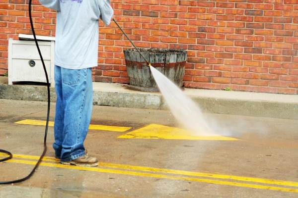 Person pressure washing pavement beside a brick wall and trash can in a parking area