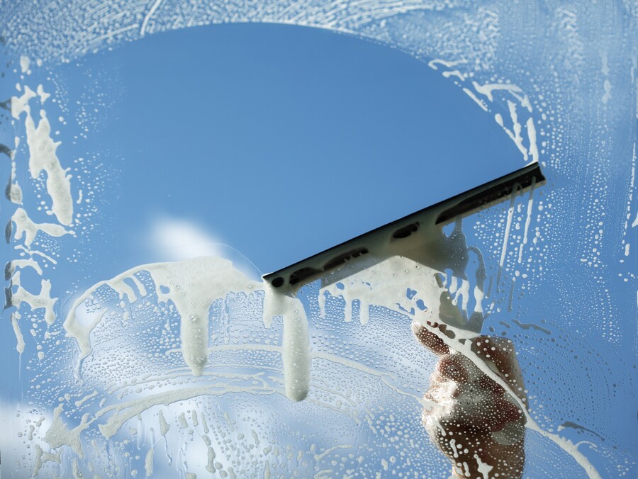 Hand using a squeegee to clean a soap-covered window against a blue sky