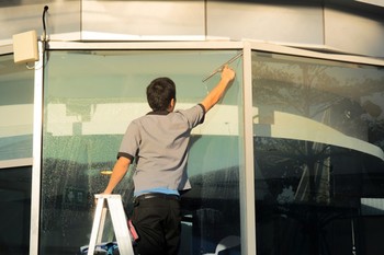 Man on a step ladder cleaning a large glass storefront window with a squeegee